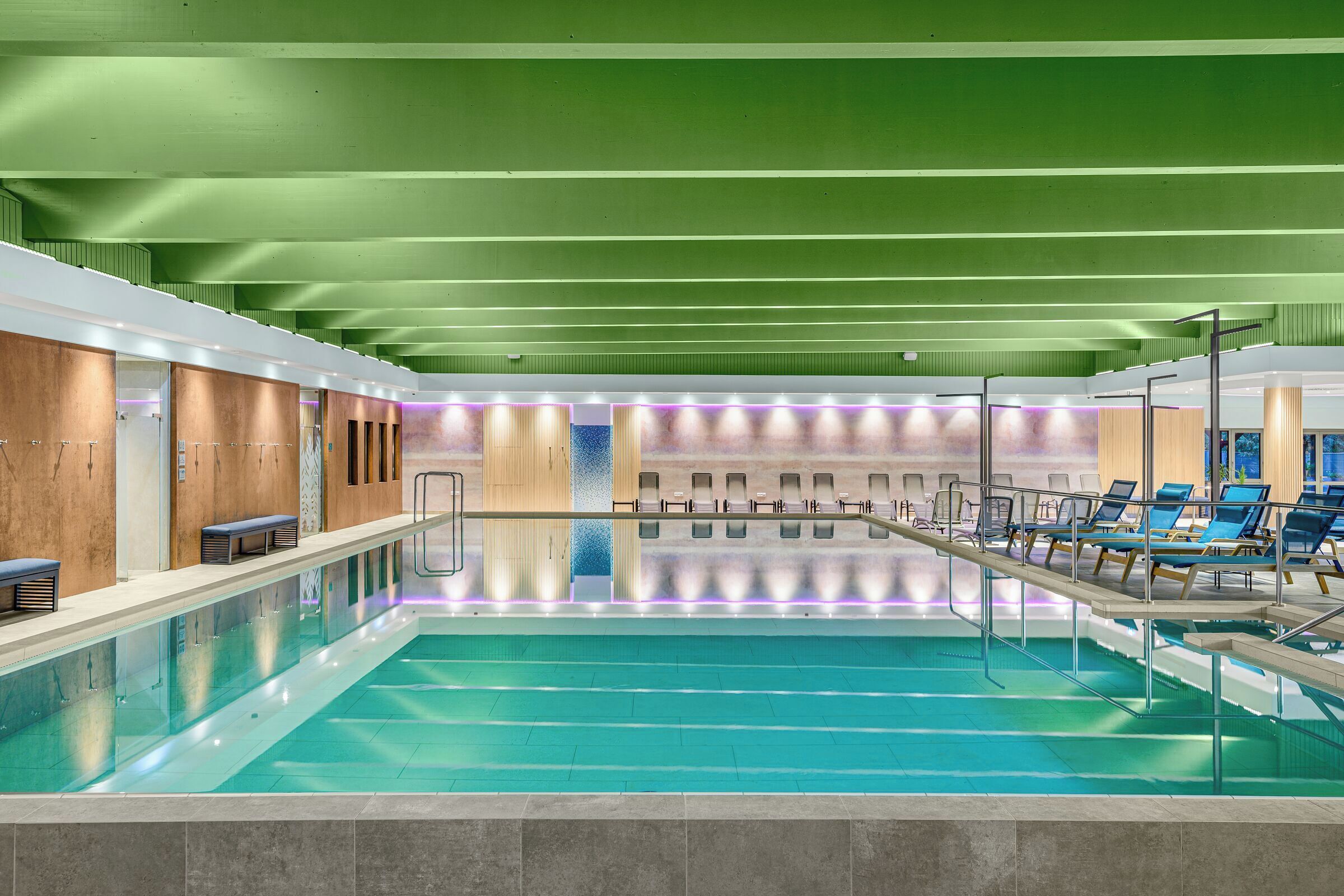 A modern indoor swimming pool with a green ceiling, lounge chairs, and a long bench along the poolside.