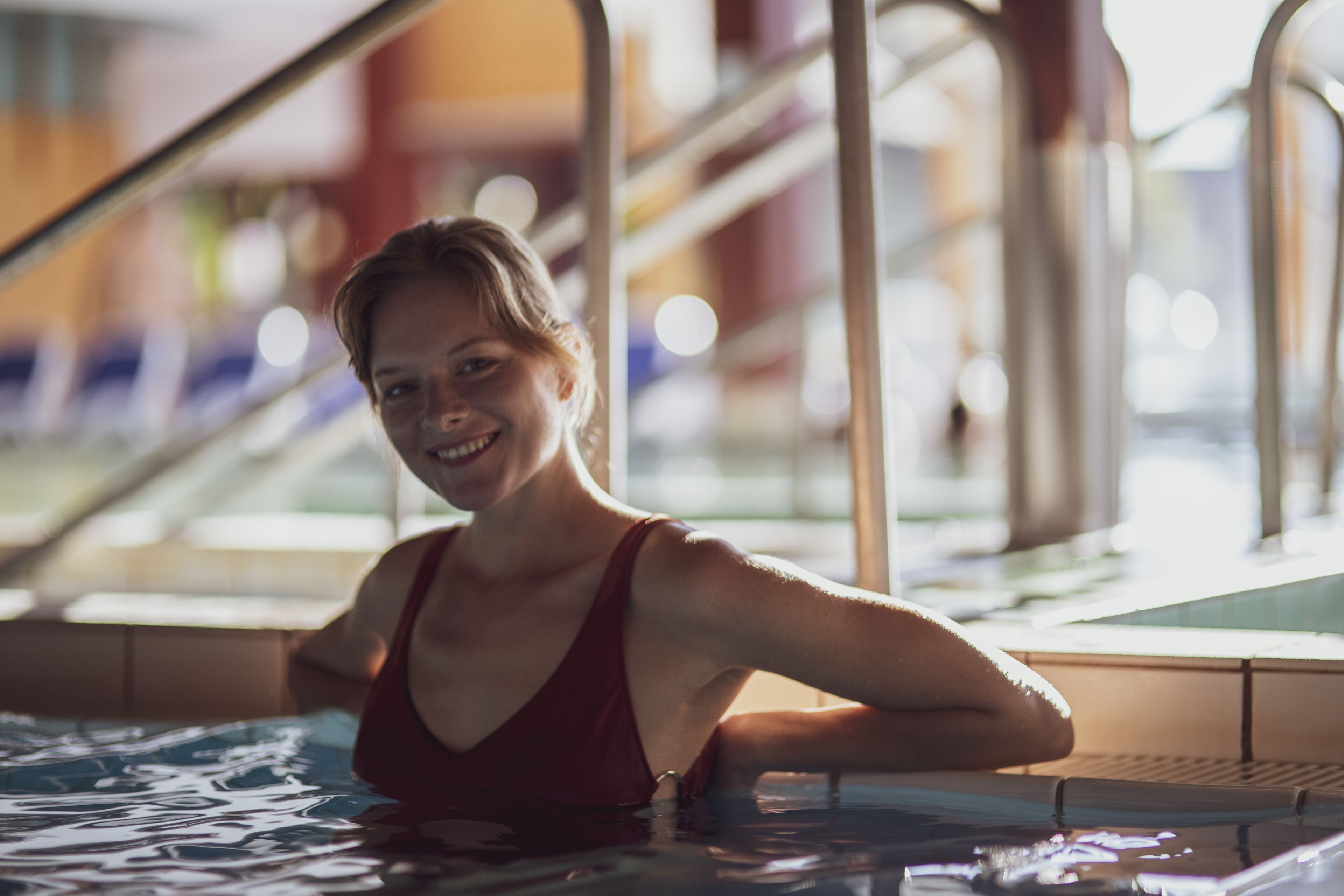 A woman in a red swimsuit in a pool area with metal railings and a tiled floor.