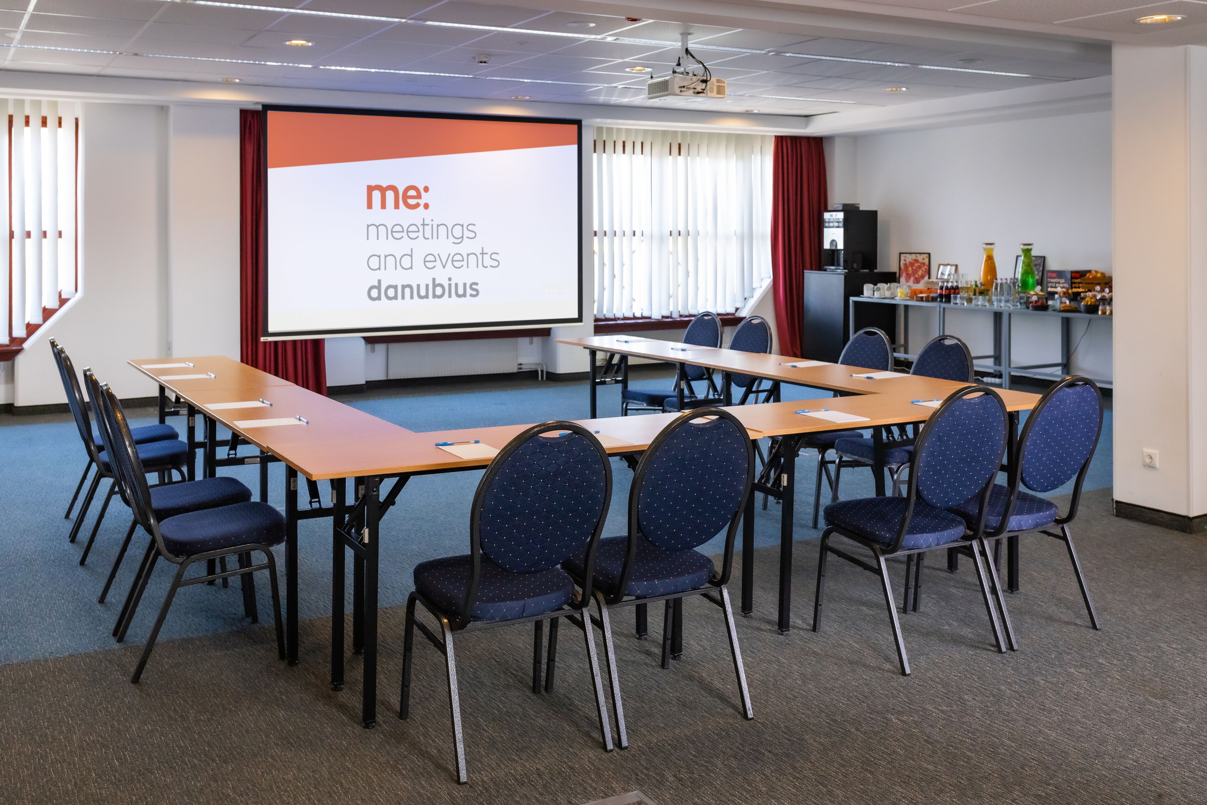 Une salle de conférence dotée d’un grand écran affichant le texte « me : réunions et événements Danubius », d’une longue table entourée de chaises et d’un petit coin bar avec des bouteilles.