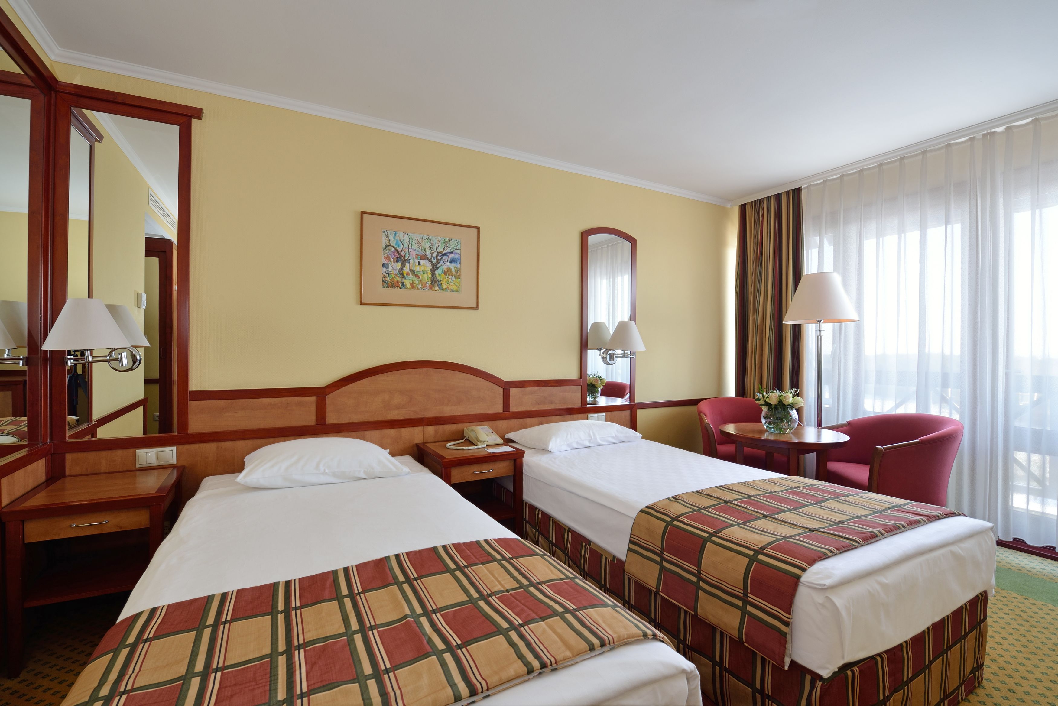Ein Hotelzimmer mit zwei Betten, einem Schreibtisch, einem Stuhl, einem Spiegel und einem Fenster mit Vorhängen.