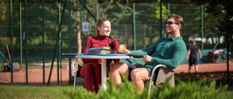 Dos personas sentadas en una mesa con una raqueta de tenis y una bebida, en un parque con una valla y árboles al fondo.