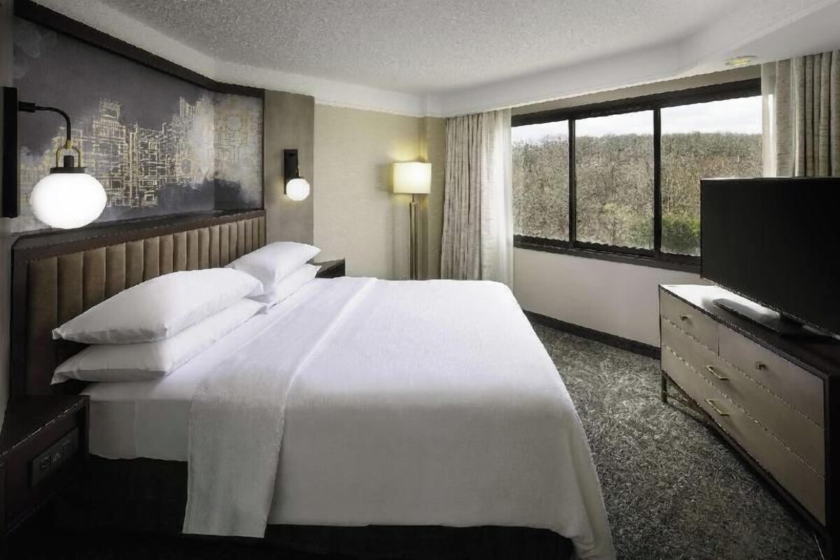 A hotel room with a bed, a TV, a cityscape painting, and a window with a view.