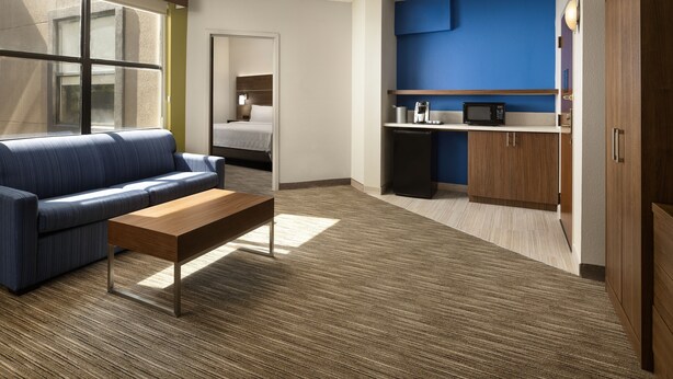 A modern hotel room with a blue sofa, a wooden coffee table, a kitchenette with a microwave, and a bed in the background.