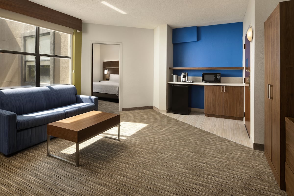 Een moderne hotelkamer met een blauwe bank, een houten bijzettafeltje, een kitchenette met een magnetron, en een bed op de achtergrond.