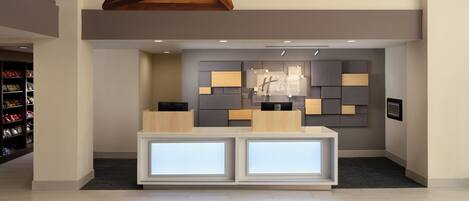 Un espace d’accueil d’hôtel au design moderne, comprenant un comptoir d’accueil, un panneau d’information fixé au mur et des luminaires suspendus aux formes géométriques.
