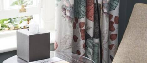 A room with a round glass table holding a magazine and a box, near a window with curtains.
