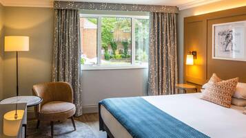Une chambre d’hôtel avec un lit, une chaise, une petite table, une lampe et une fenêtre avec des rideaux.