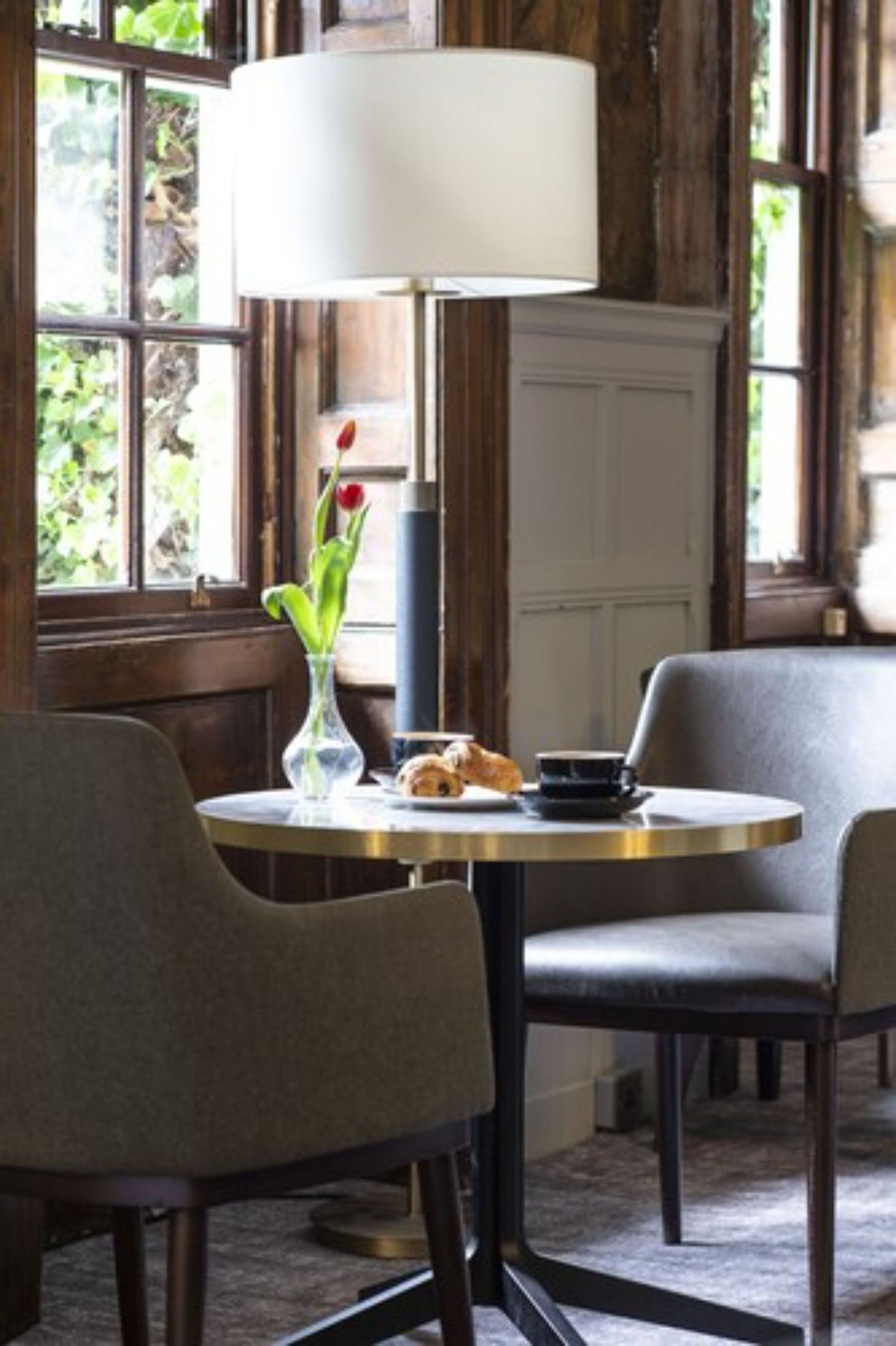 A cozy dining area with a round table, two chairs, a vase with flowers, a lamp, and a window with greenery outside.