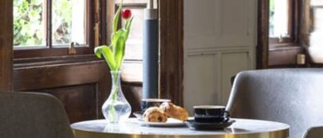 A cozy dining area with a round table, two chairs, a vase with flowers, a lamp, and a window with greenery outside.