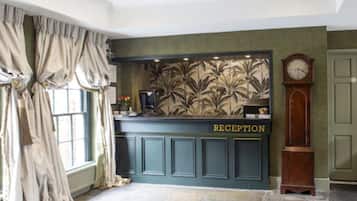 Una zona de recepción con un mostrador azul oscuro, una pared con motivos de hojas tropicales y un reloj.