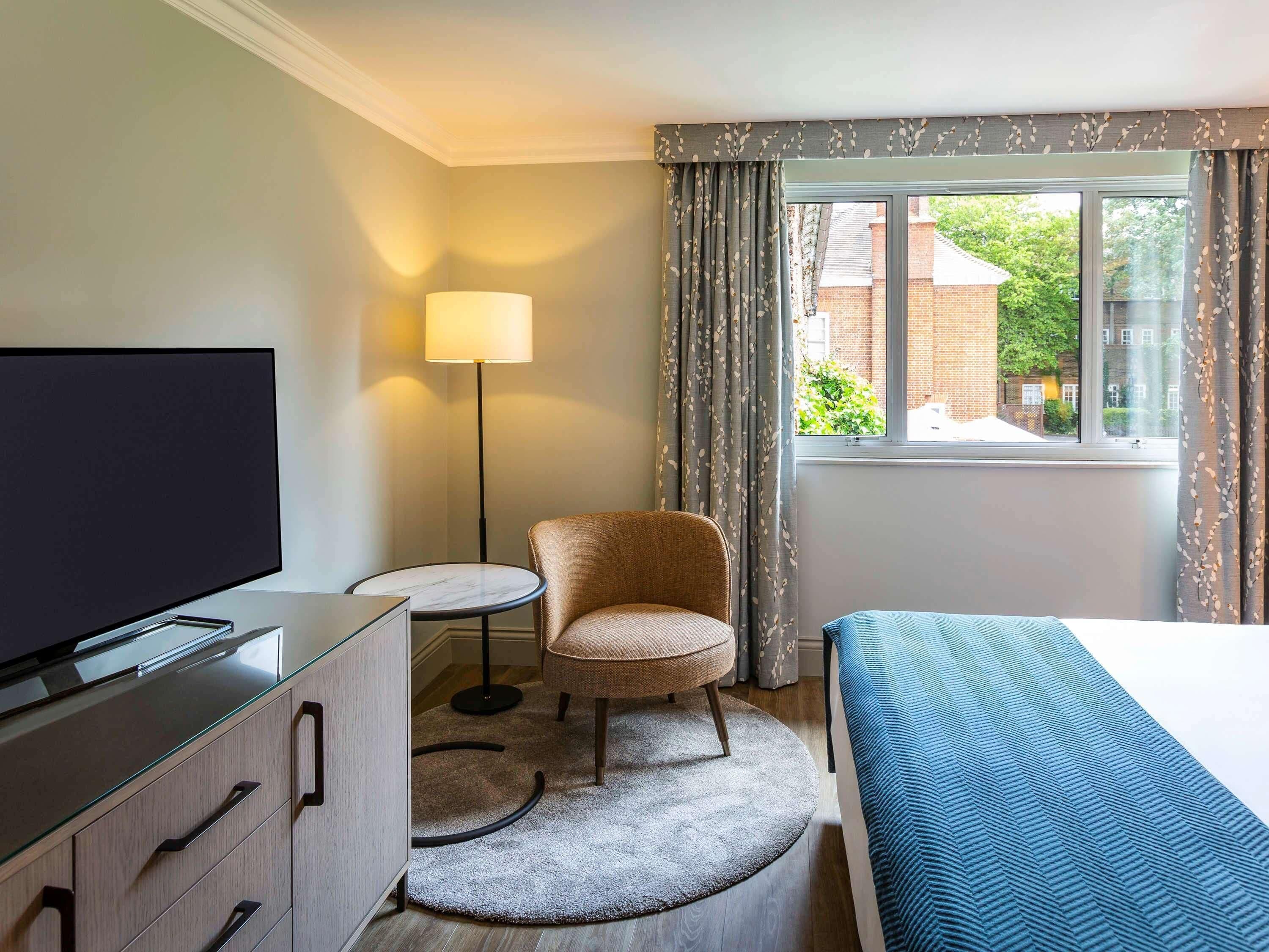 Ein Hotelzimmer mit einem Bett, einem Fernseher, einem Sessel, einer Lampe und einem Fenster mit Blick auf ein Backsteingebäude und Grünflächen.