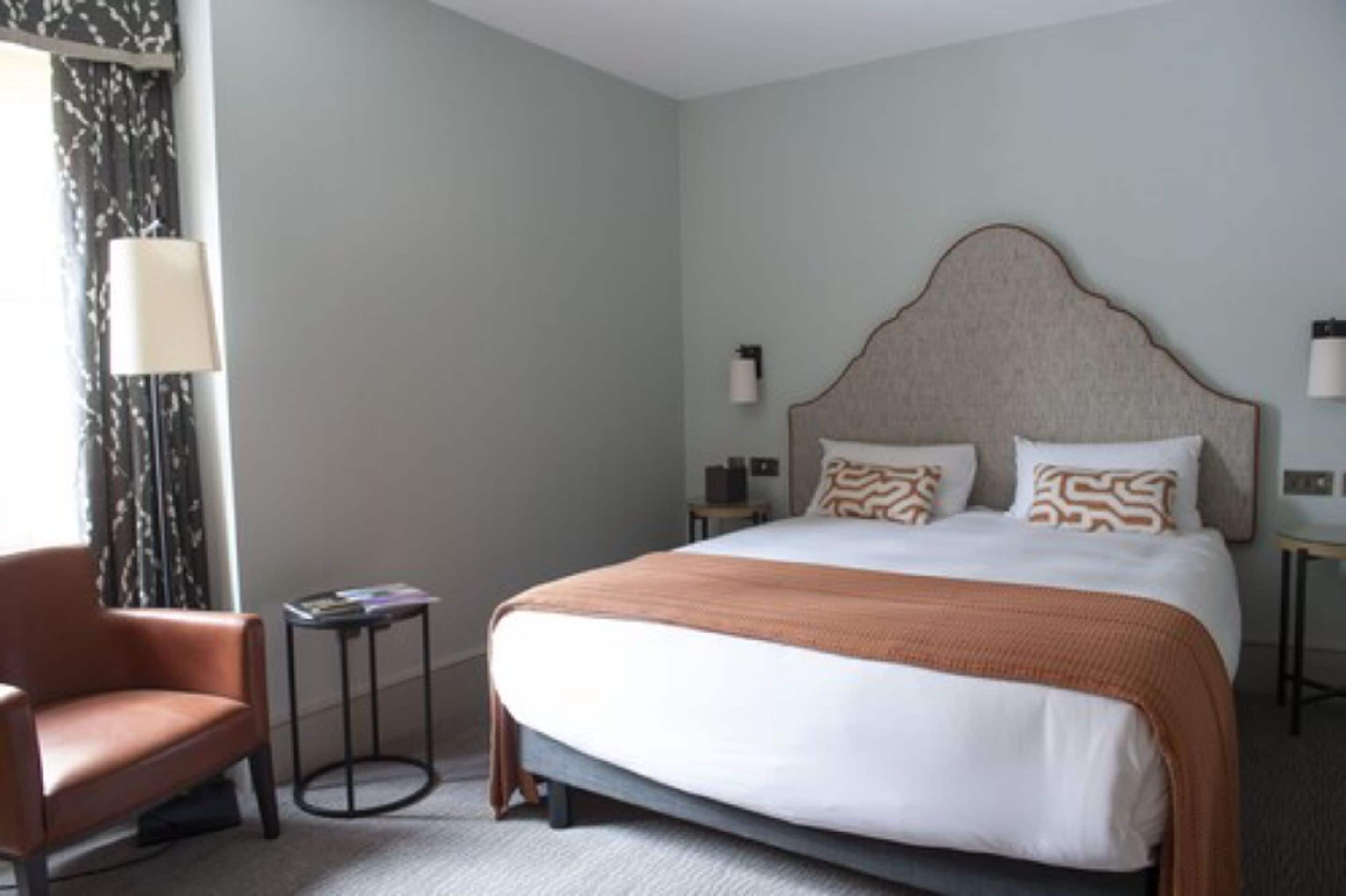 A hotel room with a large bed, a chair, a small table, and a lamp.