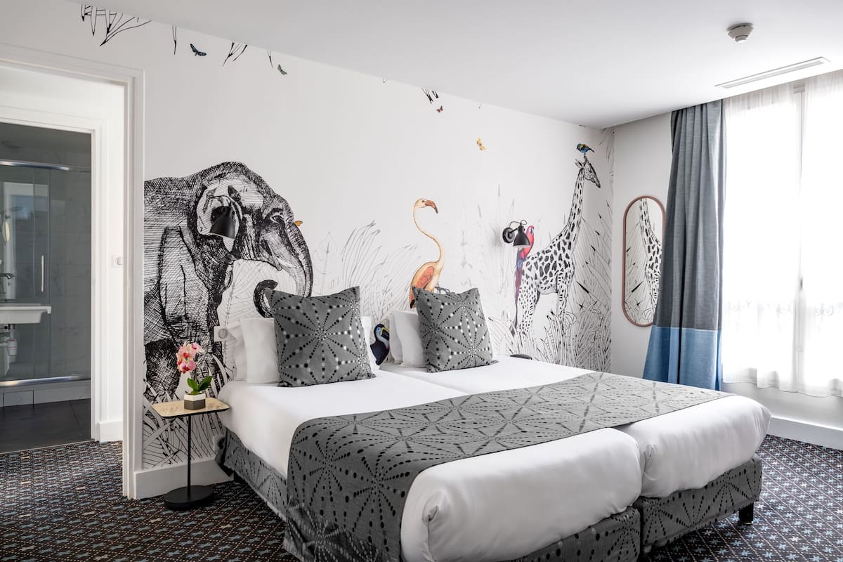Une chambre d’hôtel moderne avec un grand lit, une table de chevet ornée d’un vase et une fresque murale représentant des girafes et des oiseaux.