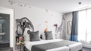 Una habitación de hotel moderna con una cama grande, una mesita de noche con un florero y un mural en la pared con jirafas y pájaros.
