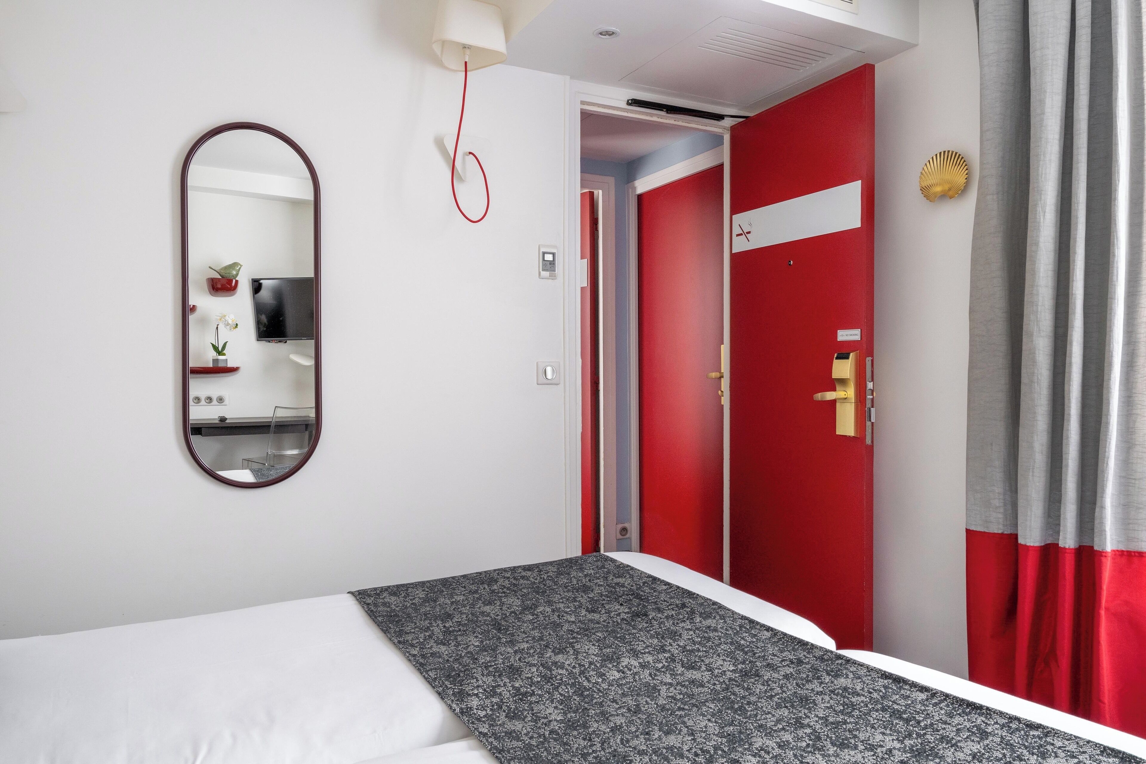 A hotel room with a bed, a red door, a mirror, and a small desk with a lamp.