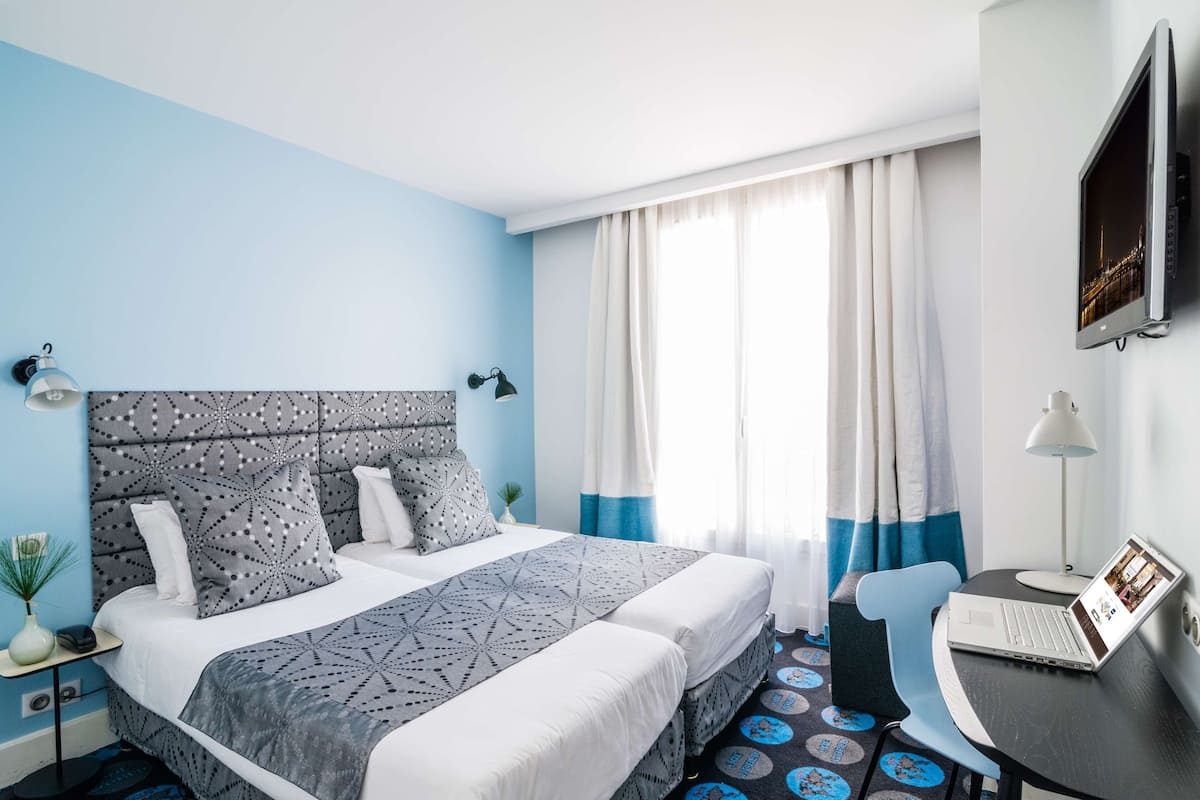 Une chambre d’hôtel avec un lit, un bureau avec un ordinateur portable, une chaise, une télévision et une fenêtre avec des rideaux.