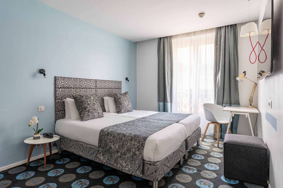 Une chambre d’hôtel avec un lit, un bureau, une chaise et une petite table sur laquelle se trouvent un téléphone et une orchidée.