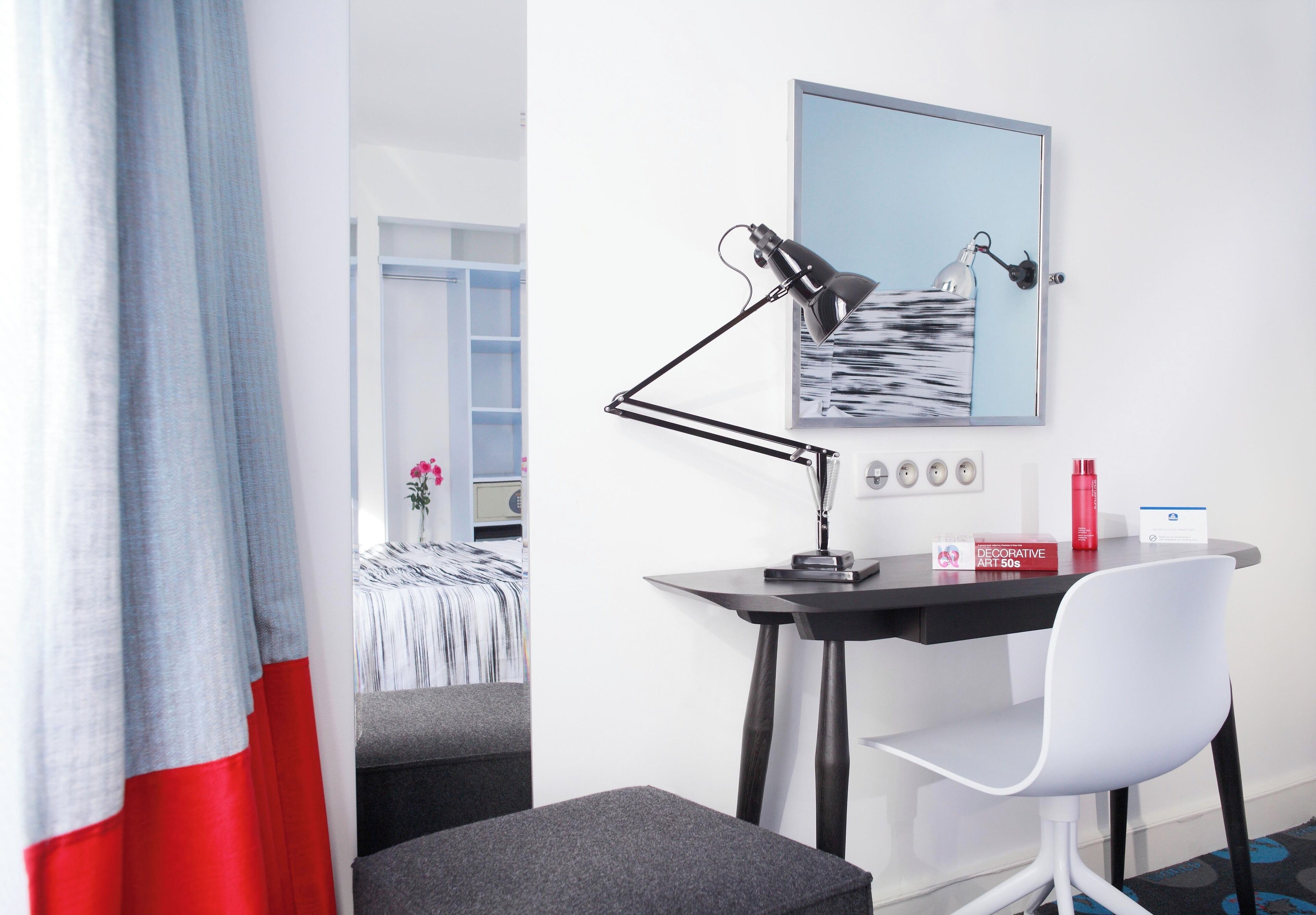 A modern hotel room with a desk, a chair, a lamp, a framed picture on the wall, and a red bottle on the desk.