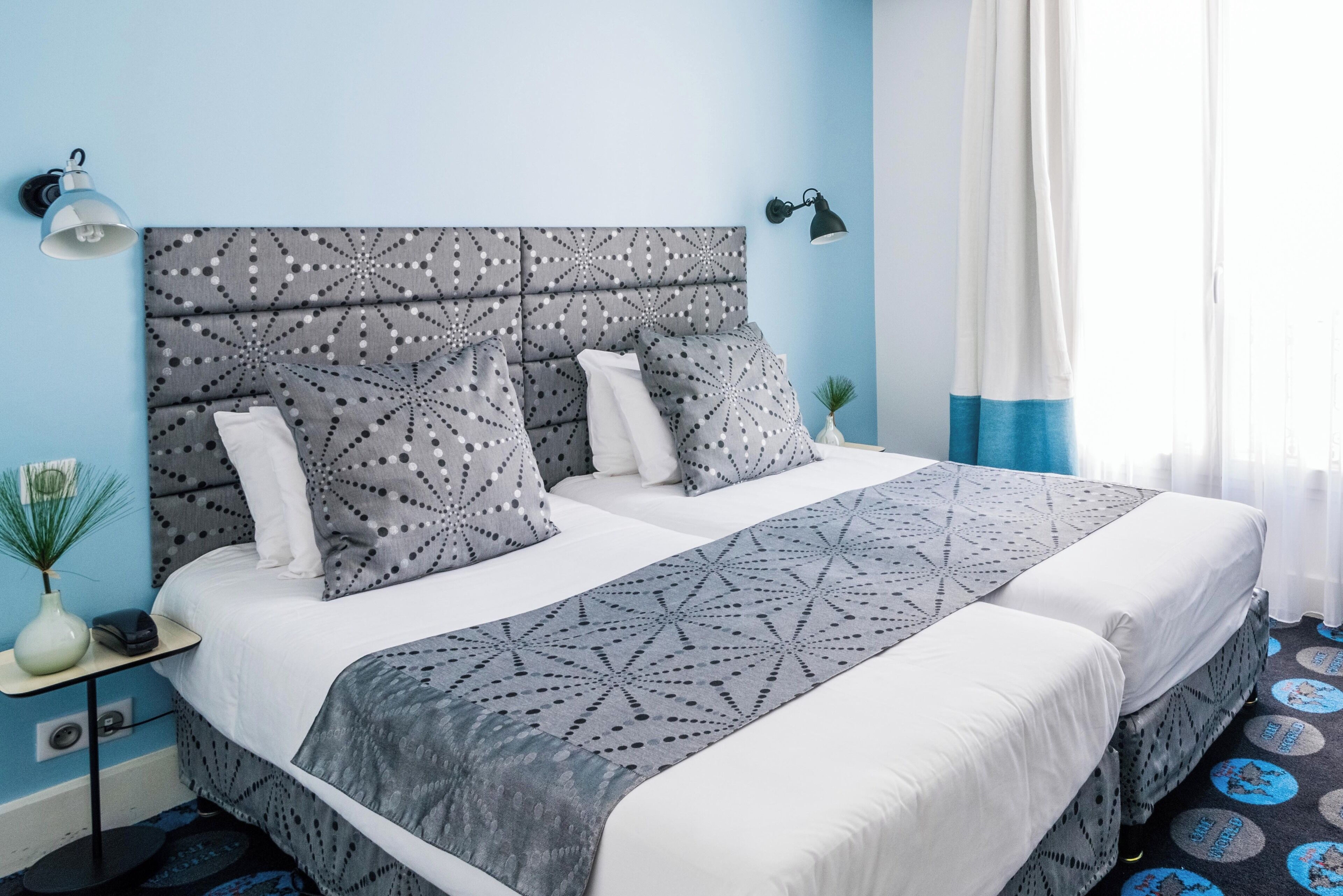 A double bed with a patterned headboard, a bedside table with a lamp, and a view of the window with curtains.