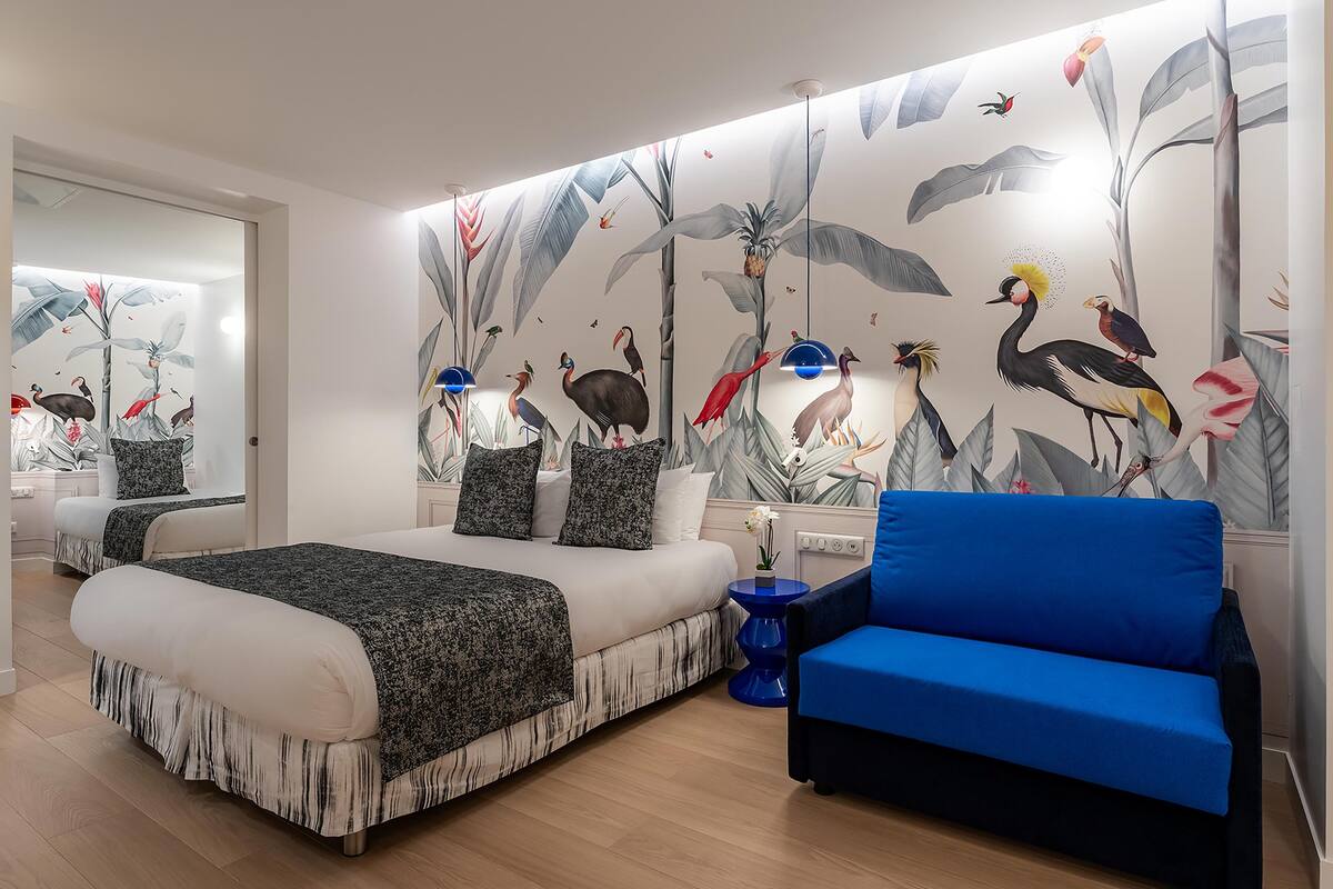 Une chambre avec un grand lit, un canapé bleu et une fresque murale représentant des oiseaux et du feuillage.