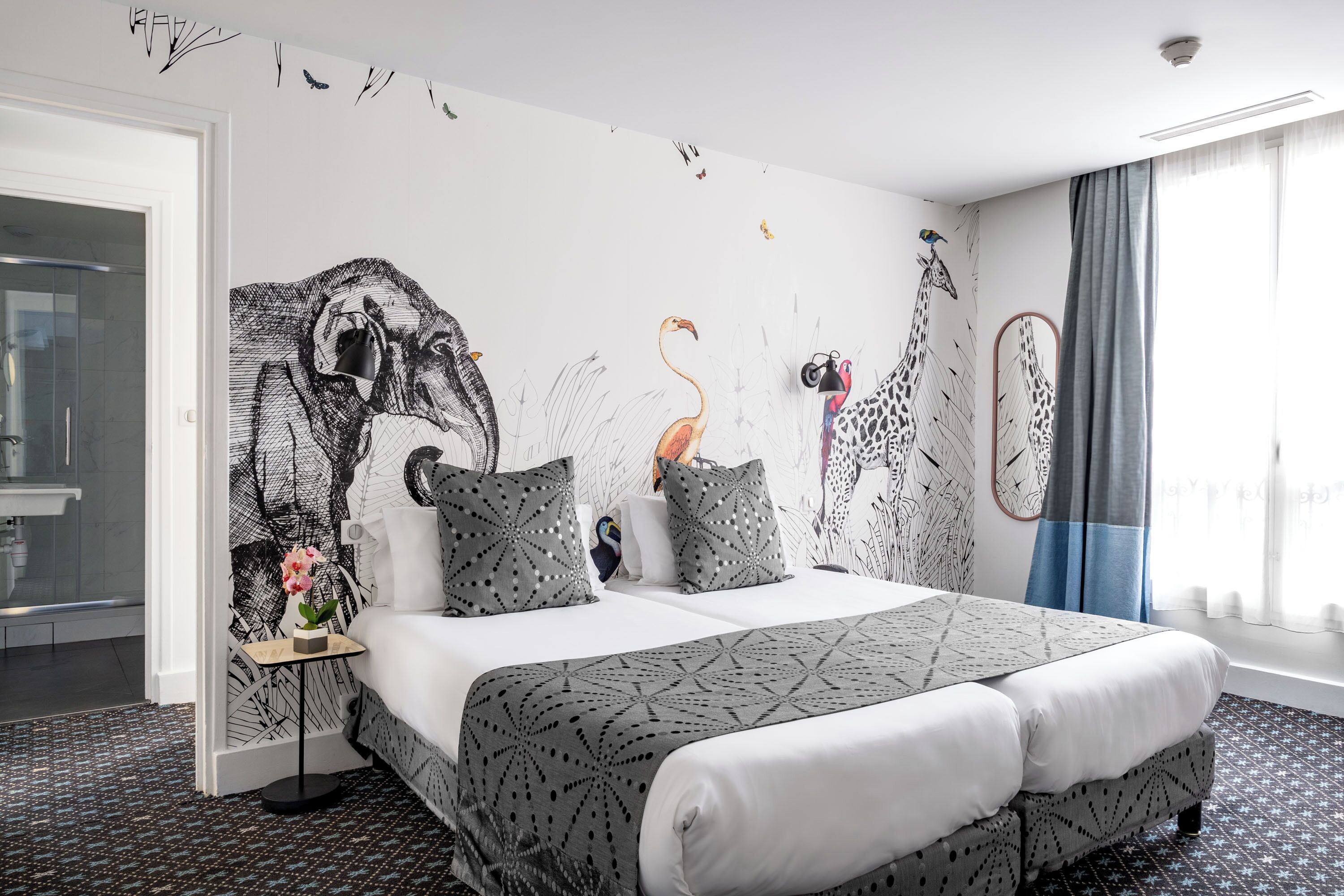 A modern hotel room with a large bed, a side table with a vase, and a wall mural of giraffes and birds.