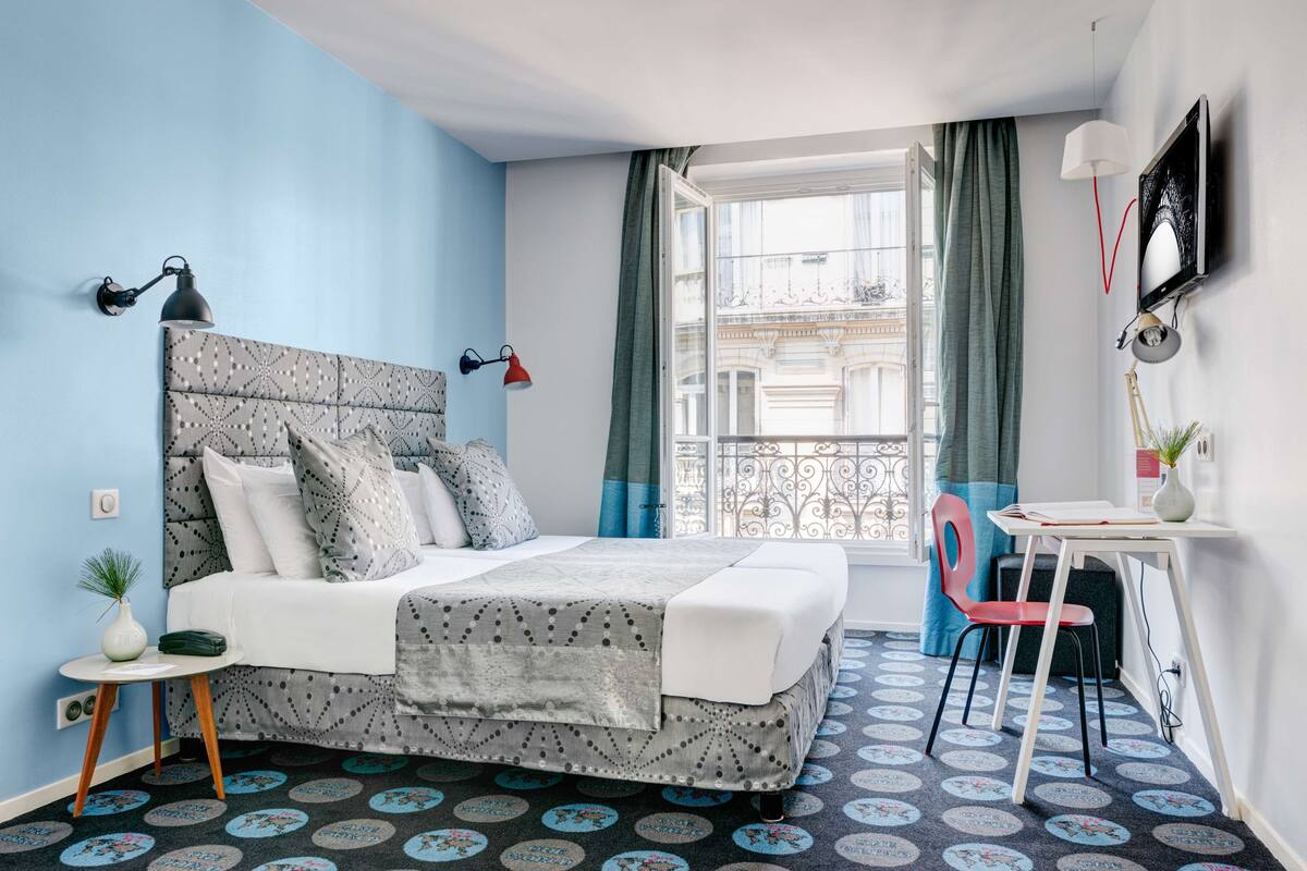 Une chambre à coucher comprenant un lit, un bureau, une chaise, une fenêtre avec des rideaux et une vue sur des immeubles.