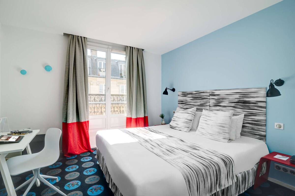 Une chambre d’hôtel avec un lit, un bureau, une chaise et une fenêtre avec des rideaux.