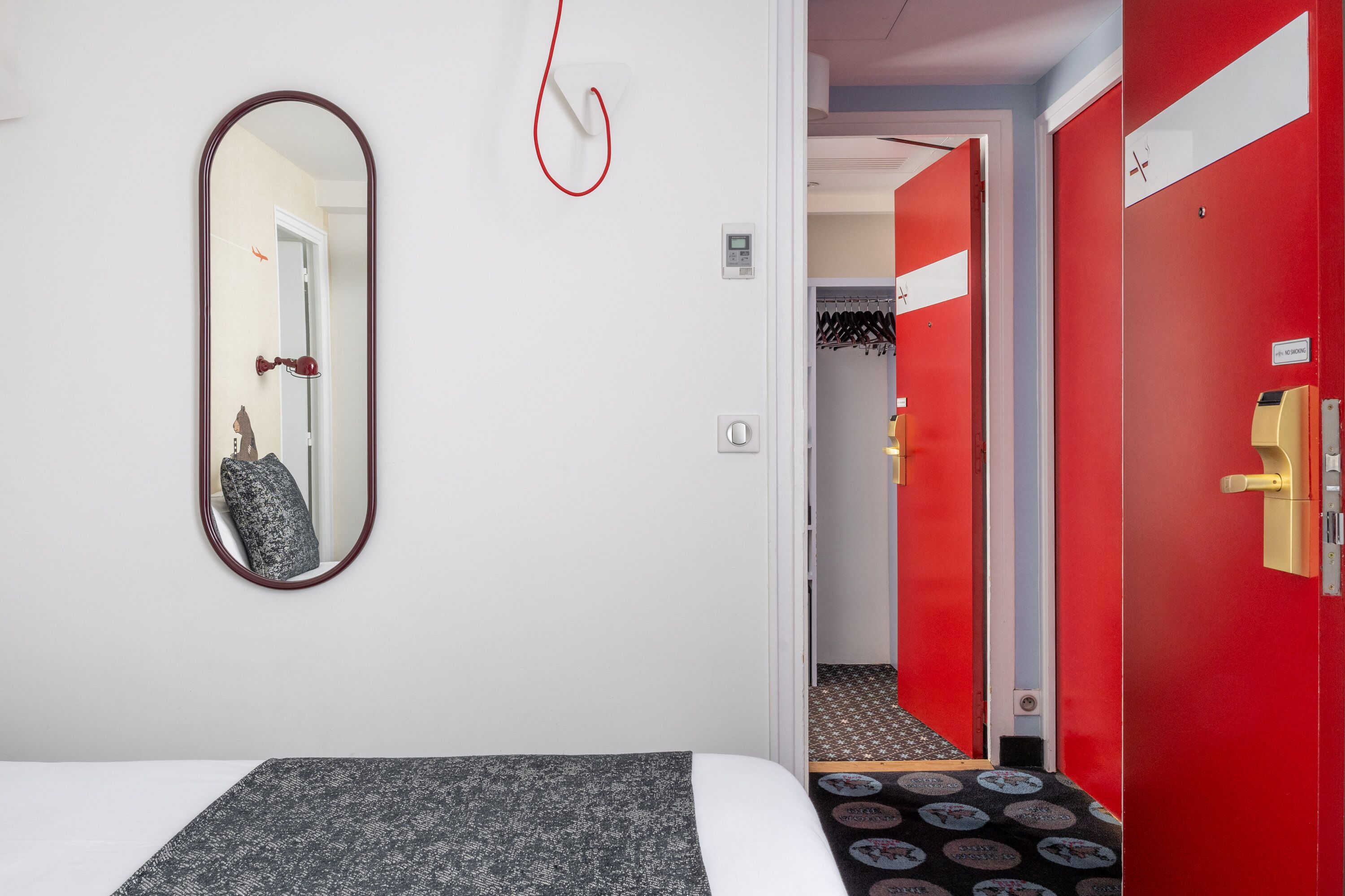 A hotel room with a bed, a mirror, a red door, and a hallway with a coat rack.