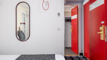 A hotel room with a bed, a mirror, a red door, and a hallway with a coat rack.