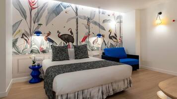 Une chambre avec un grand lit, une chaise bleue, une fresque murale représentant des oiseaux et des plantes, et un luminaire moderne.