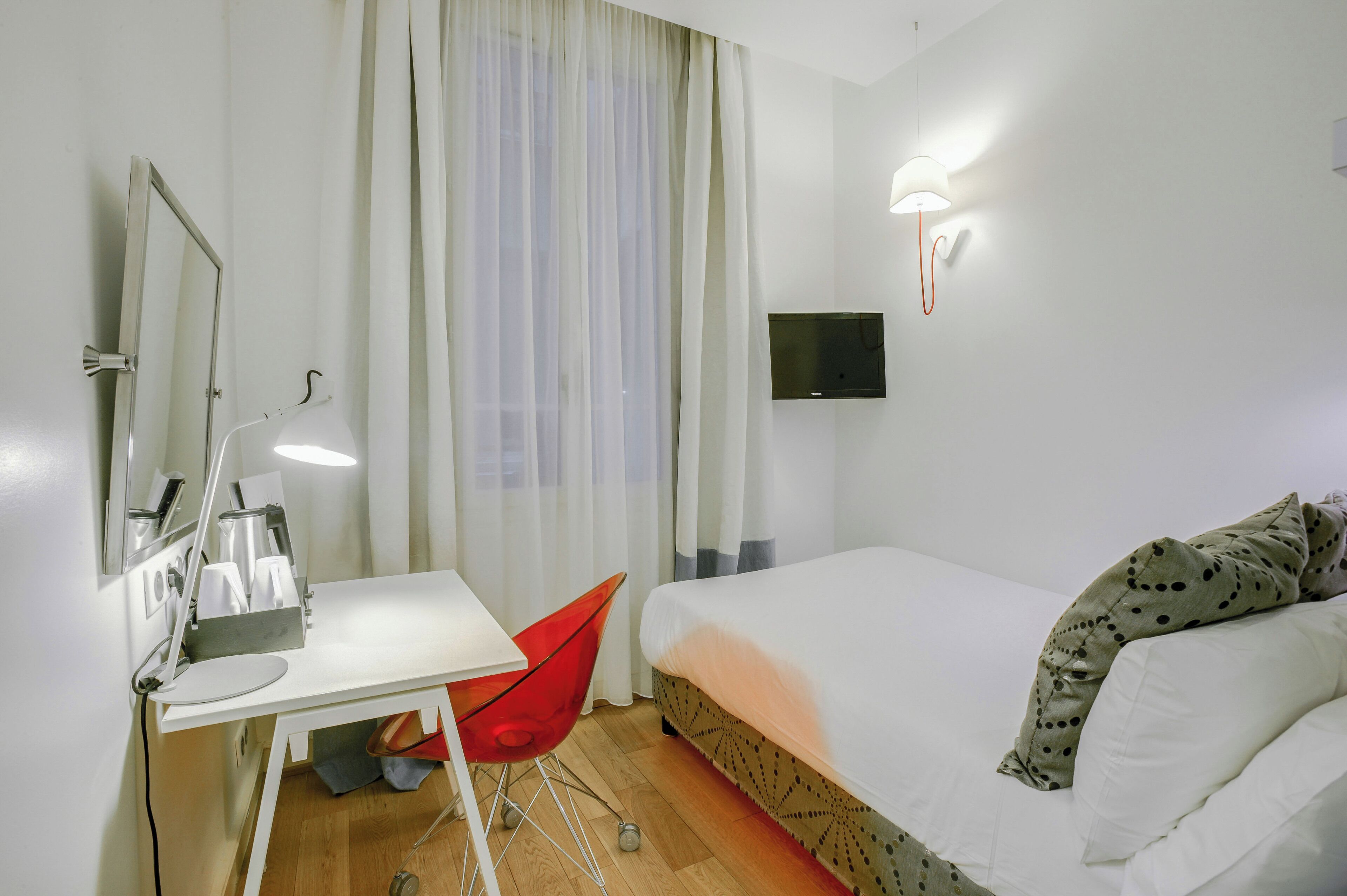 A hotel room with a bed, a desk with a chair, a mirror, and a TV mounted on the wall.