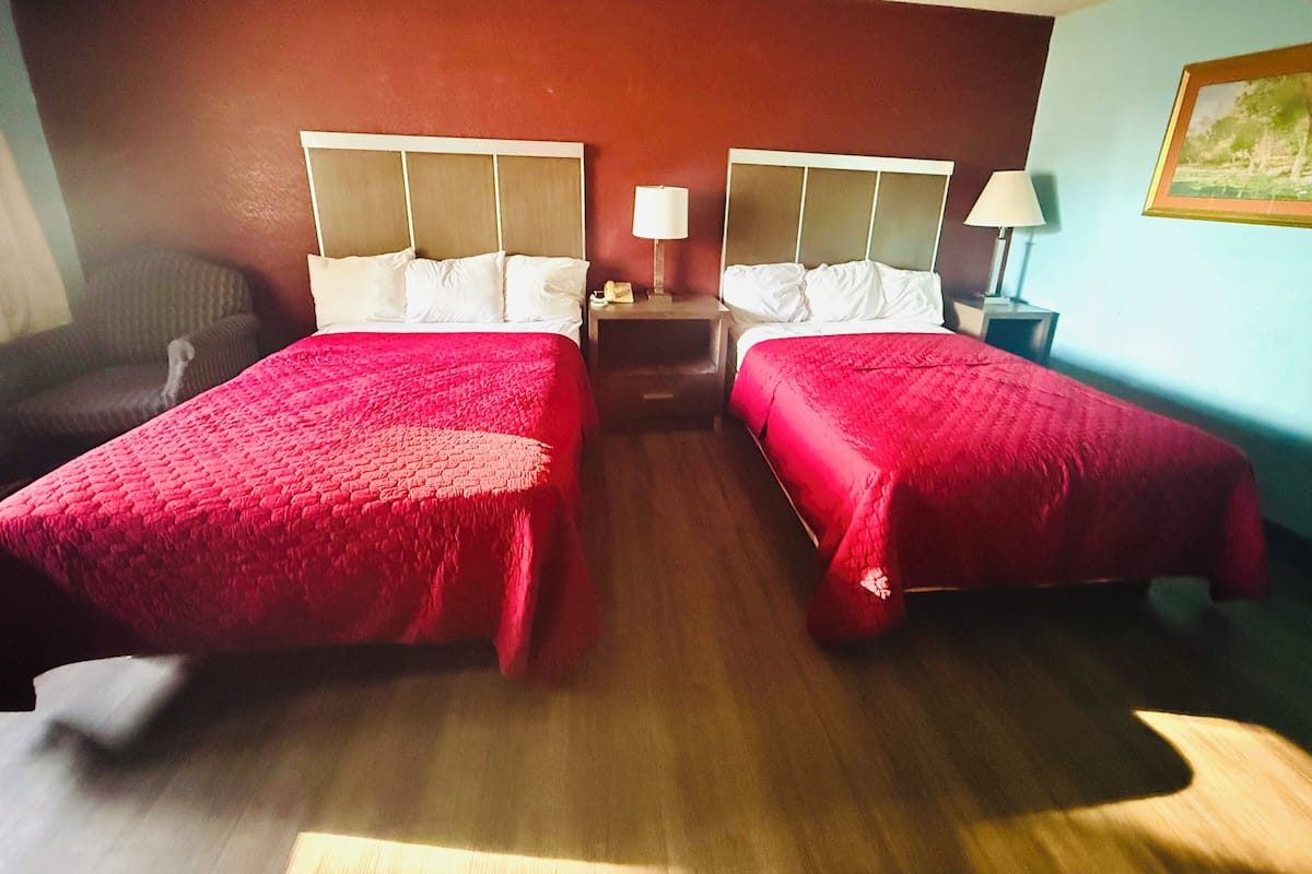 A hotel room with two beds, each with a red bedspread, wooden headboards, and white pillows. There is a nightstand with a lamp between the beds, and a painting on the wall.