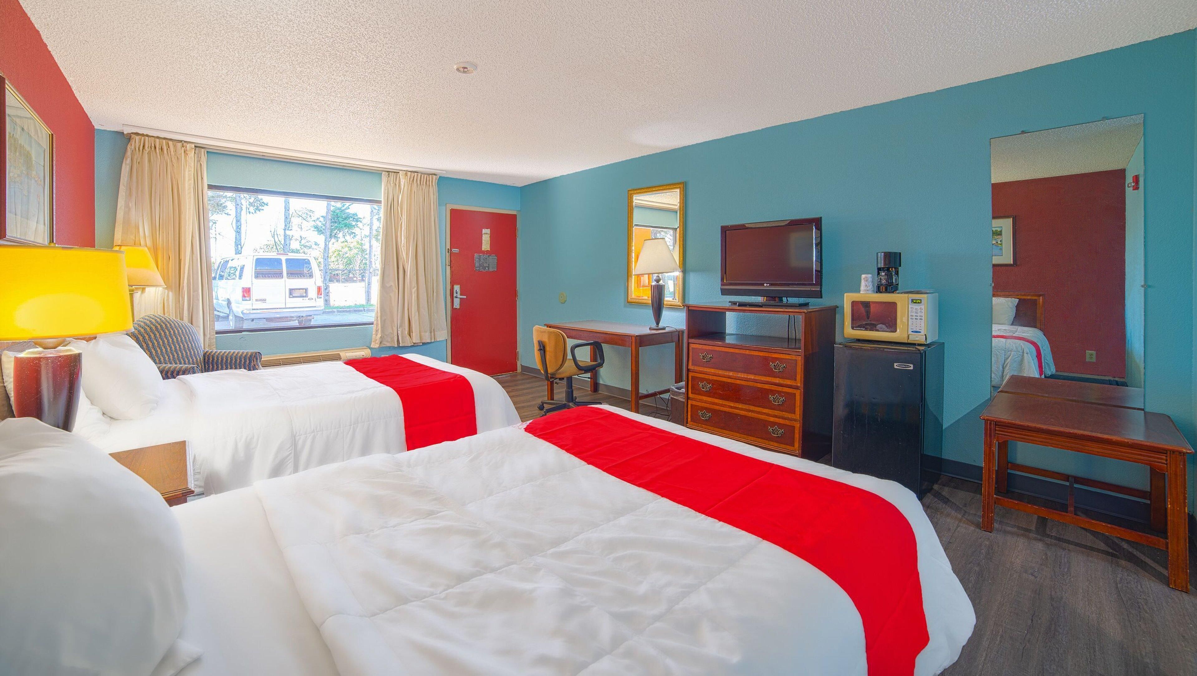 A hotel room with two beds, a desk, a television, a refrigerator, and a window with curtains.