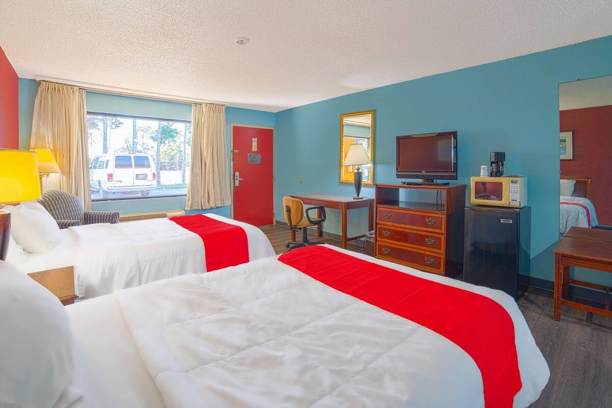 A hotel room with two beds, a desk, a television, a refrigerator, and a window with curtains.
