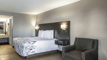 A hotel room with a bed, a chair, a nightstand, a lamp, and a bathroom with a mirror and sink.