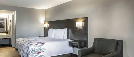 Une chambre d’hôtel comprenant un lit, une chaise, une table de chevet, une lampe et une salle de bain avec un miroir et un lavabo.