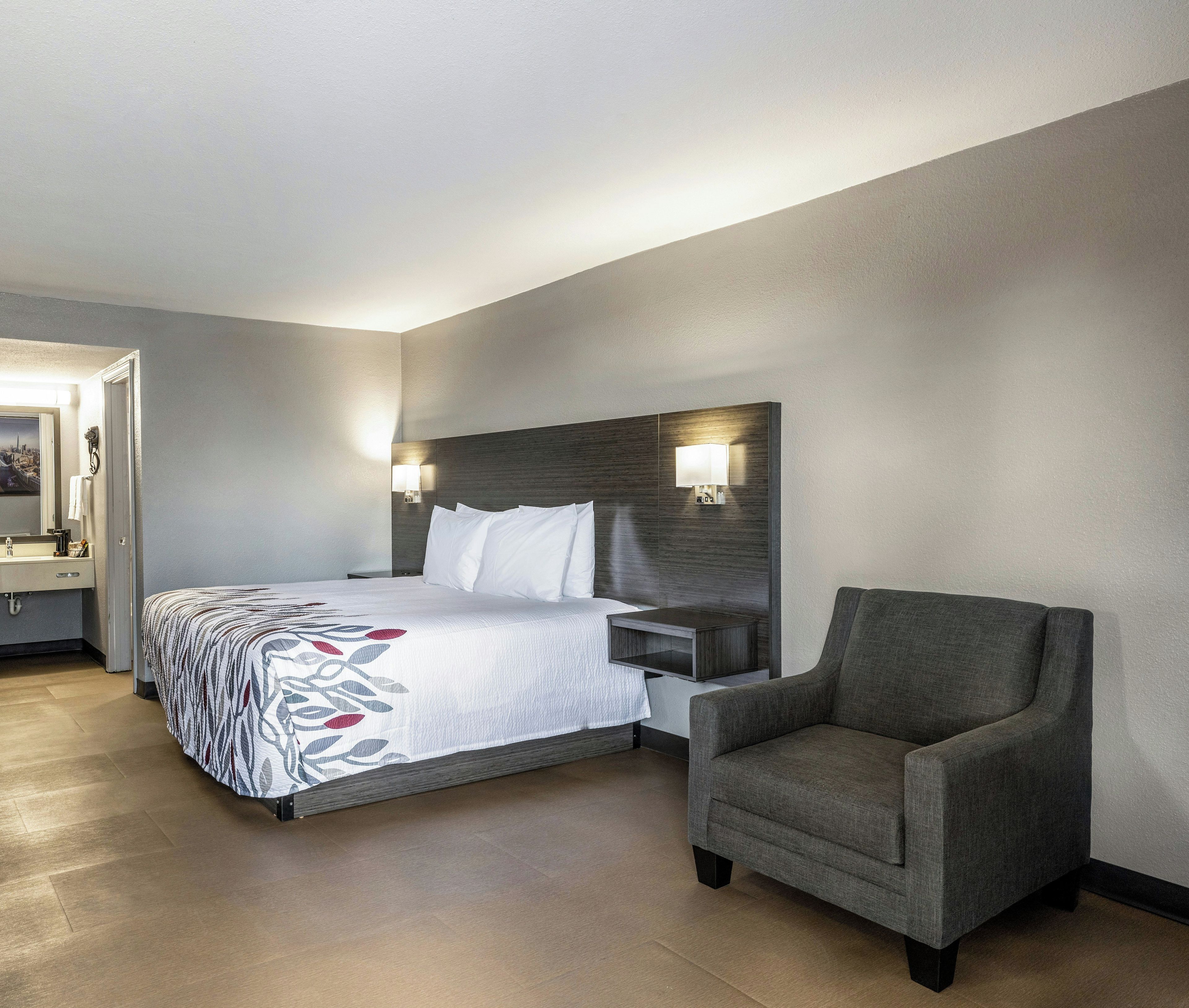 A hotel room with a bed, a nightstand, a chair, a bedside table, a lamp, and a bathroom with a mirror.