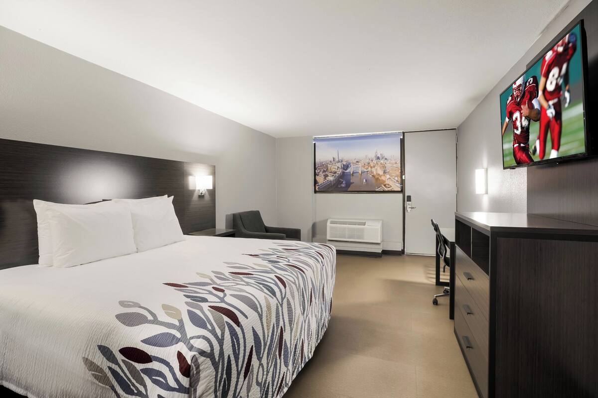 A hotel room with a large bed, a desk, a chair, a TV mounted on the wall, and an air conditioning unit.