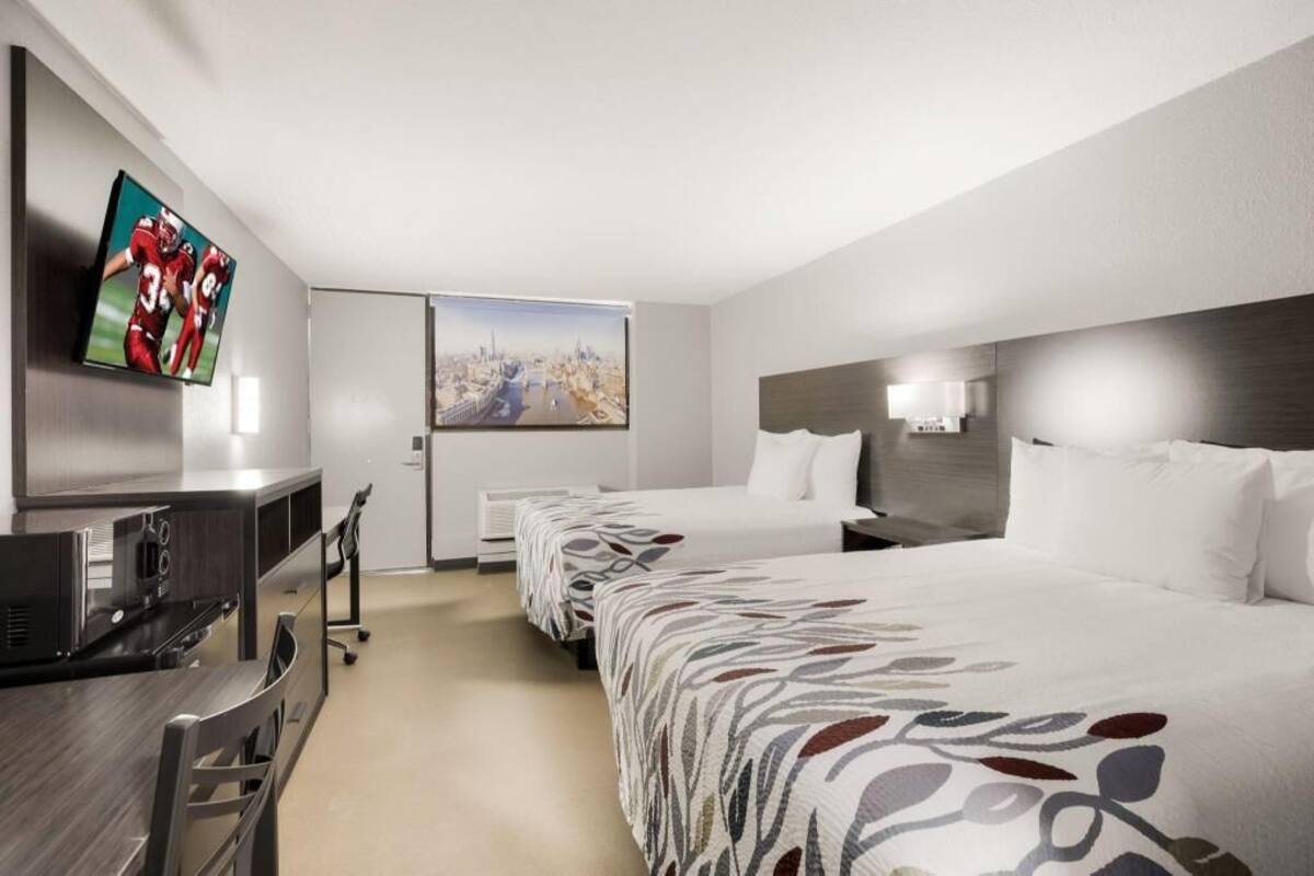 A hotel room with a TV, a desk, a bed, and a cityscape painting.