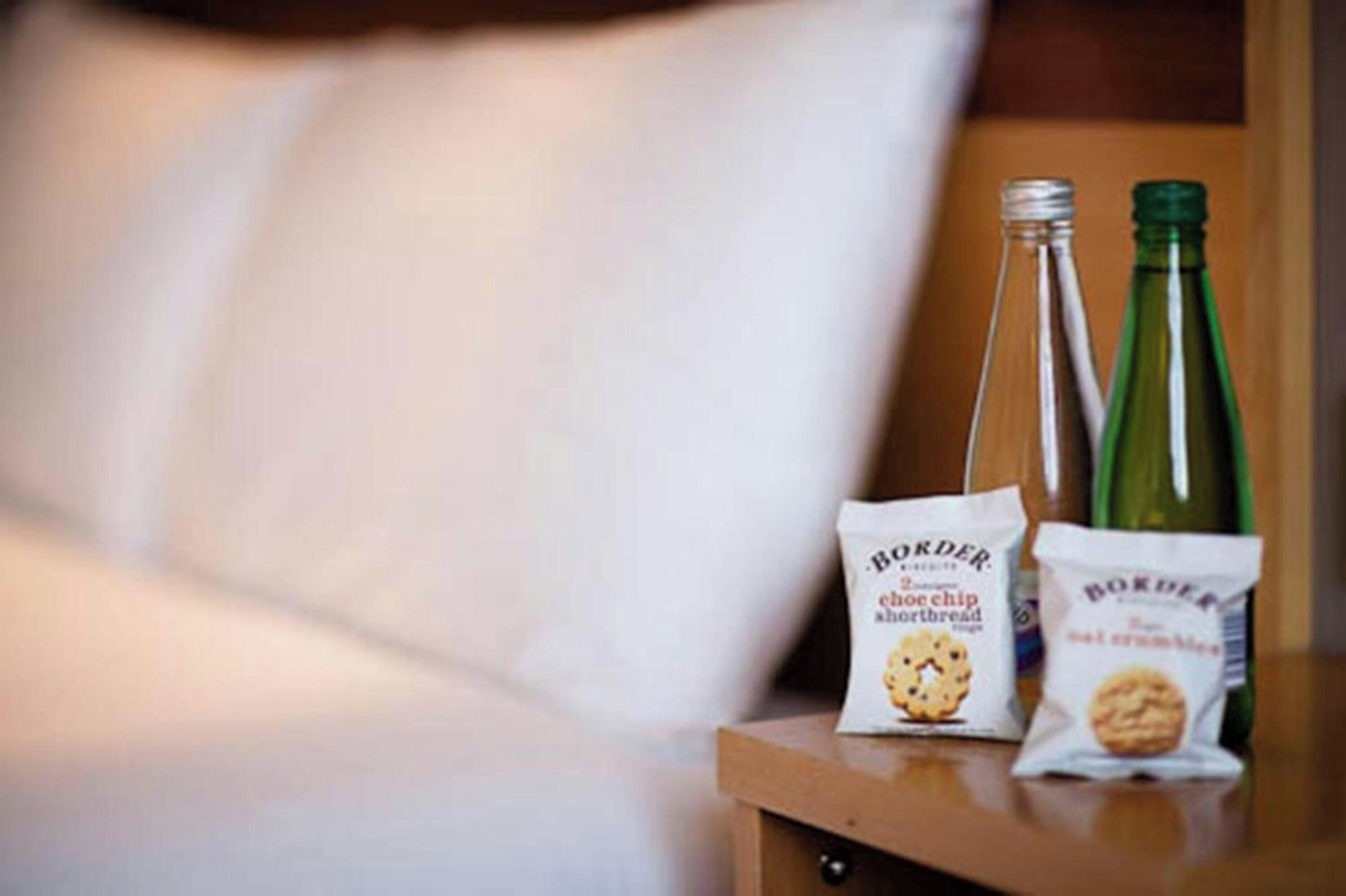 Una mesita de noche de madera con dos botellas verdes y dos paquetes de galletas chip de chocolate Border.