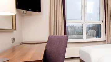 Une chambre d’hôtel avec un bureau en bois, une chaise violette, un téléviseur à écran plat fixé au mur, un lit et une fenêtre avec des rideaux.