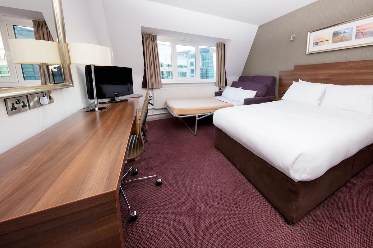 A hotel room with a bed, a desk with a TV, a chair, and a window with curtains.