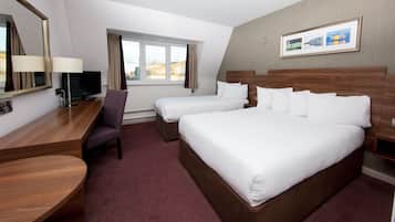 A hotel room with a large bed, two smaller beds, a desk with a chair, a TV, and a window with curtains.