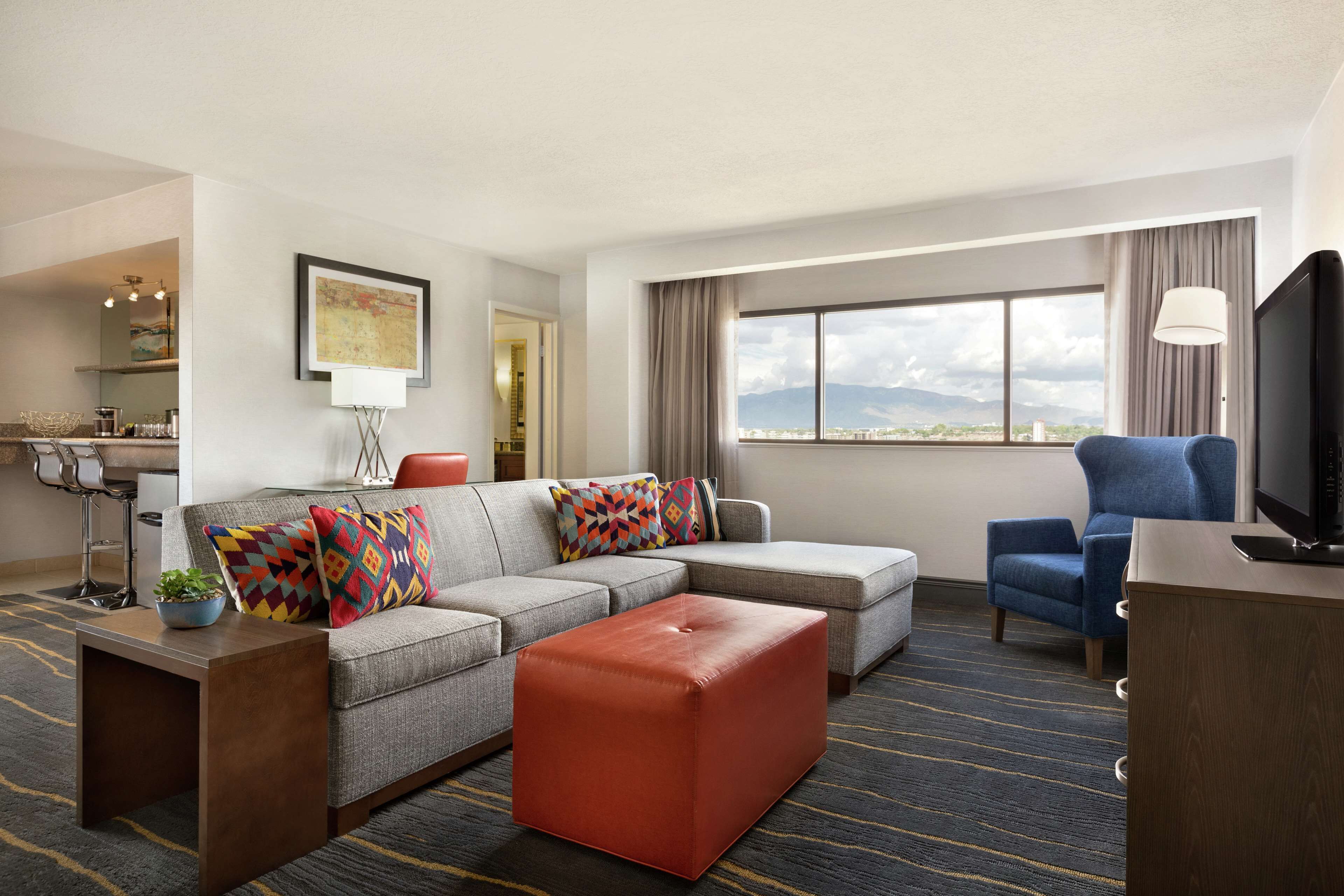 A modern hotel room with a sectional sofa, a blue armchair, a flat-screen TV, and a large window offering a city view.