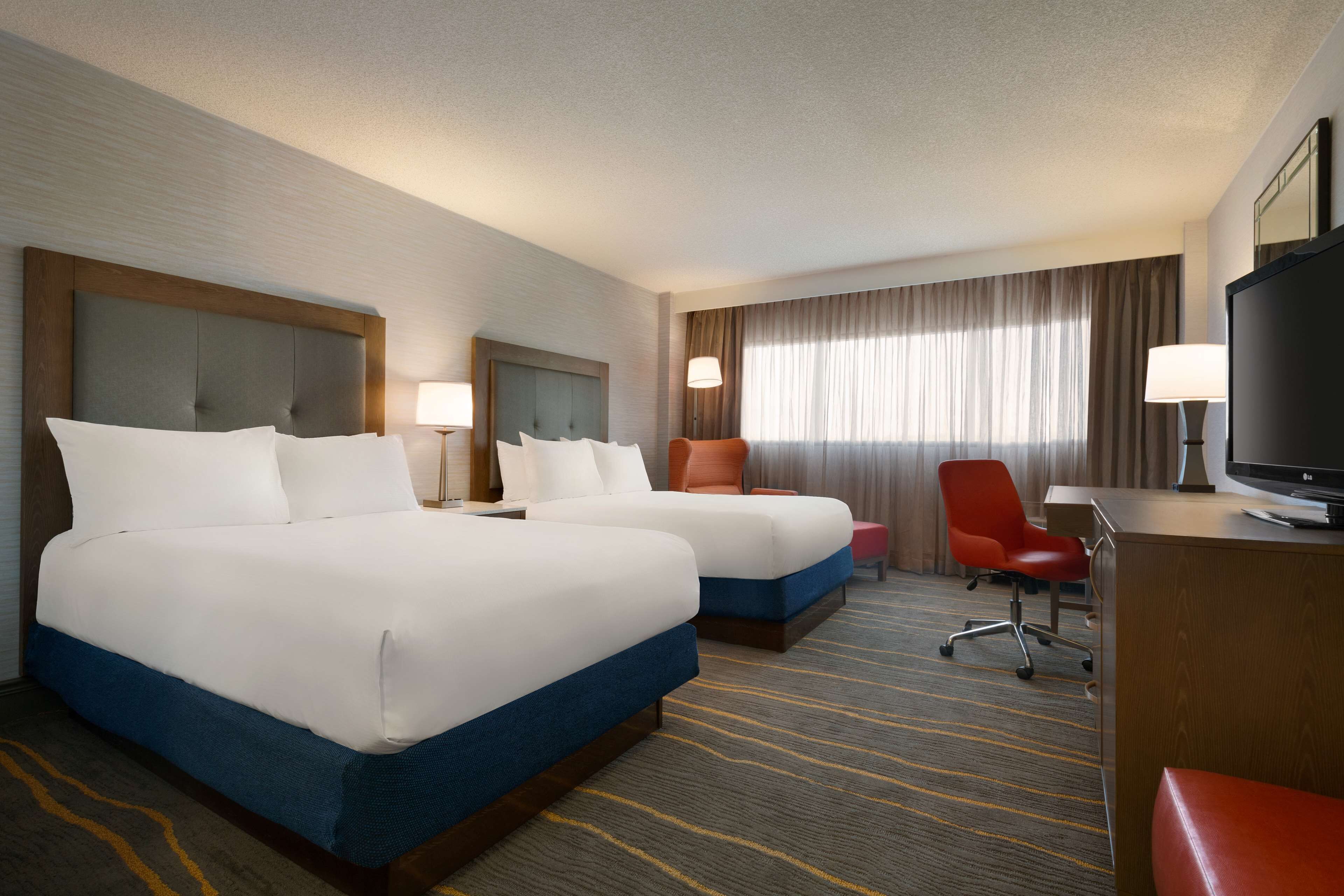 A hotel room with two beds, a desk with a TV, a red chair, and a large window with curtains.