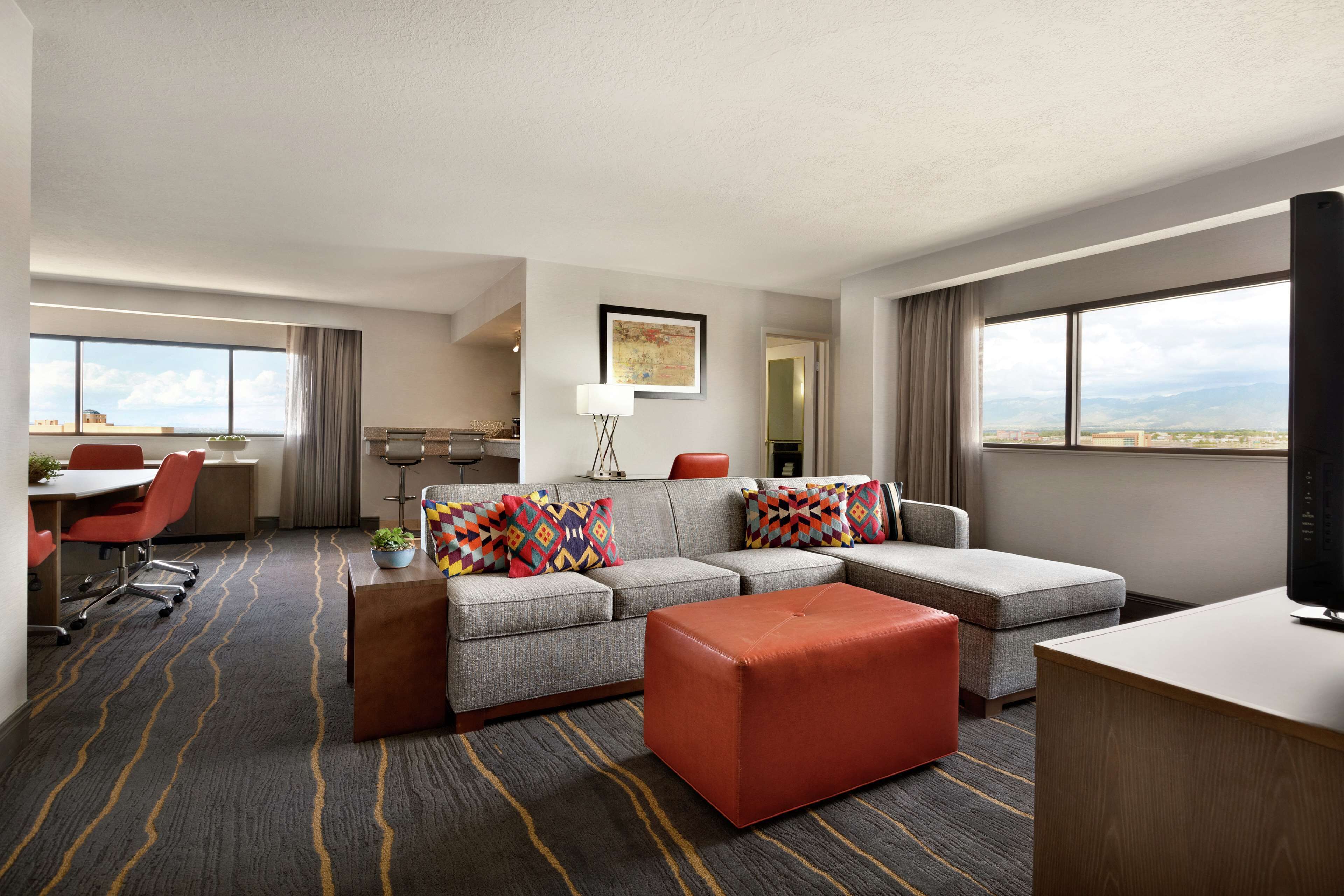 A hotel room with a grey sofa, red ottoman, a desk, a chair, and a TV.