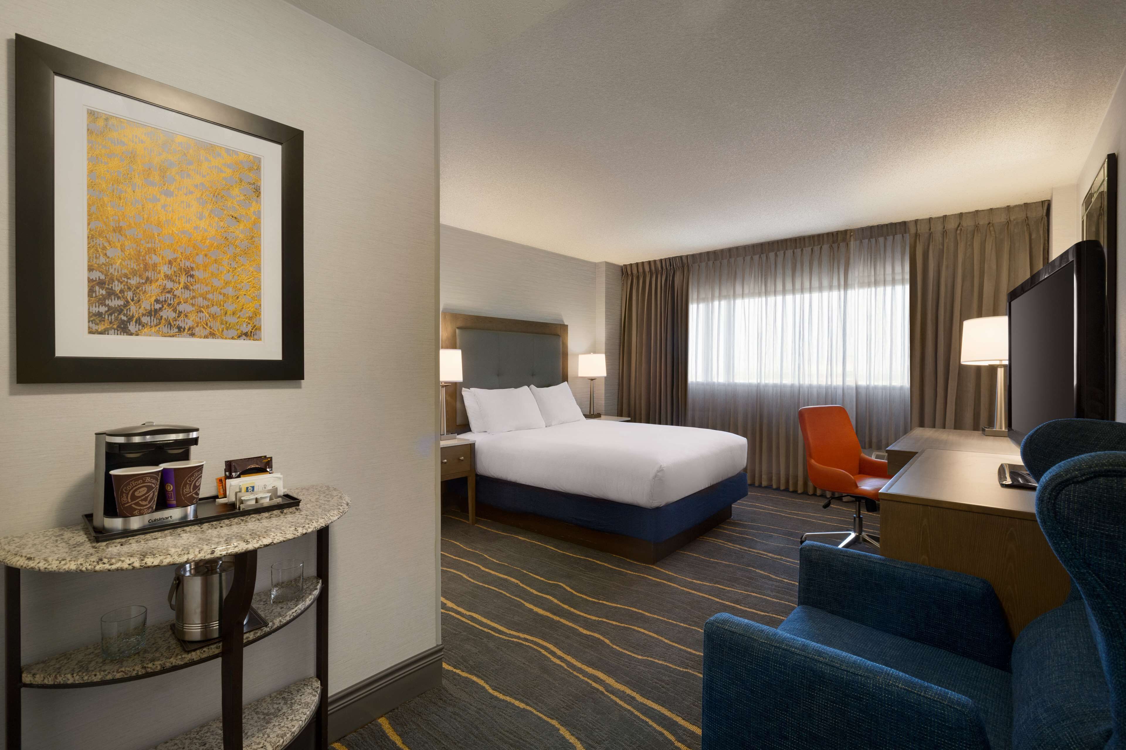 Quarto de hotel com uma cama, escrivaninha, cadeira, cafeteira e um quadro na parede.