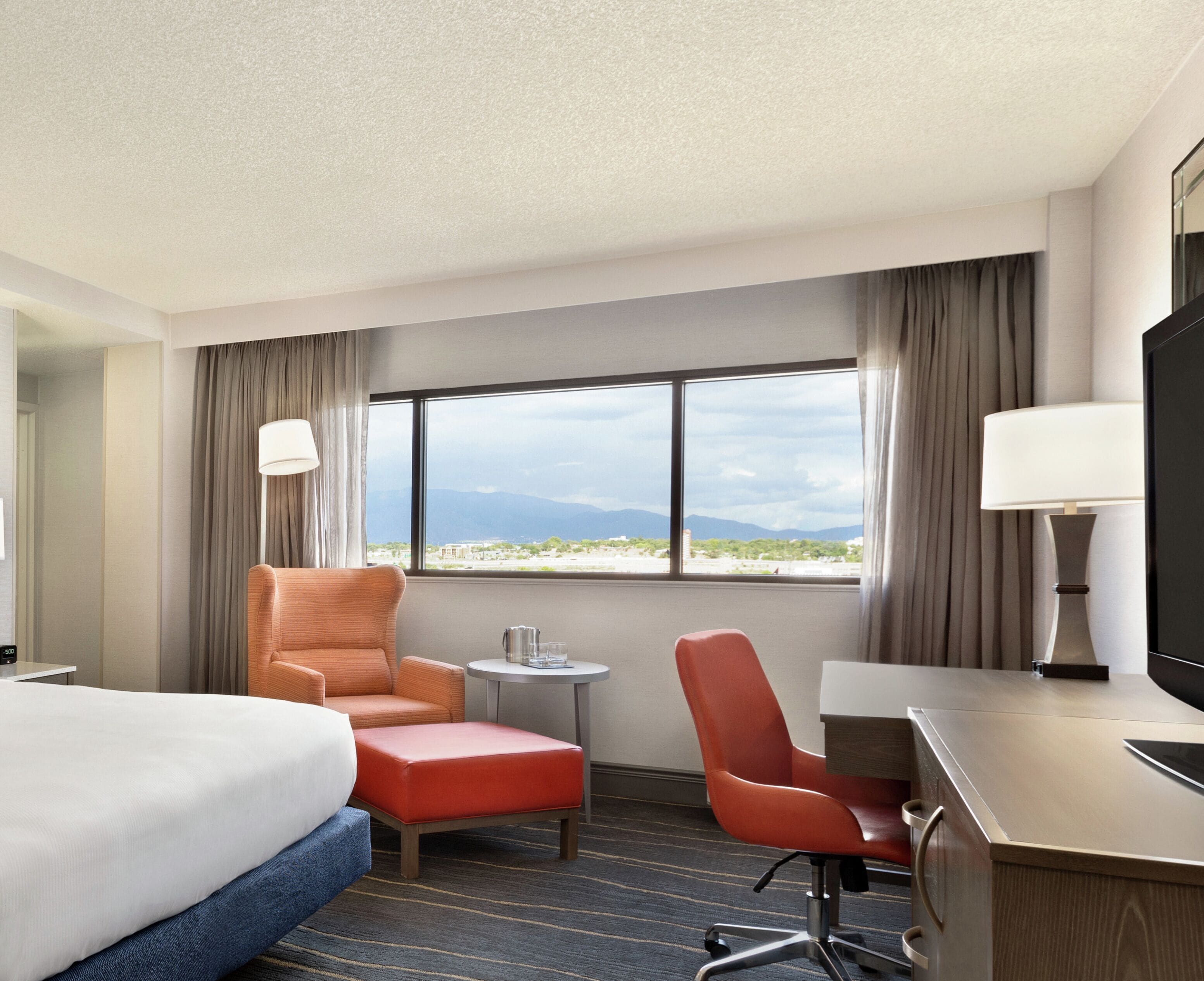 A hotel room with a bed, two orange chairs, a small table, a desk, a TV, and a window with a view of mountains.