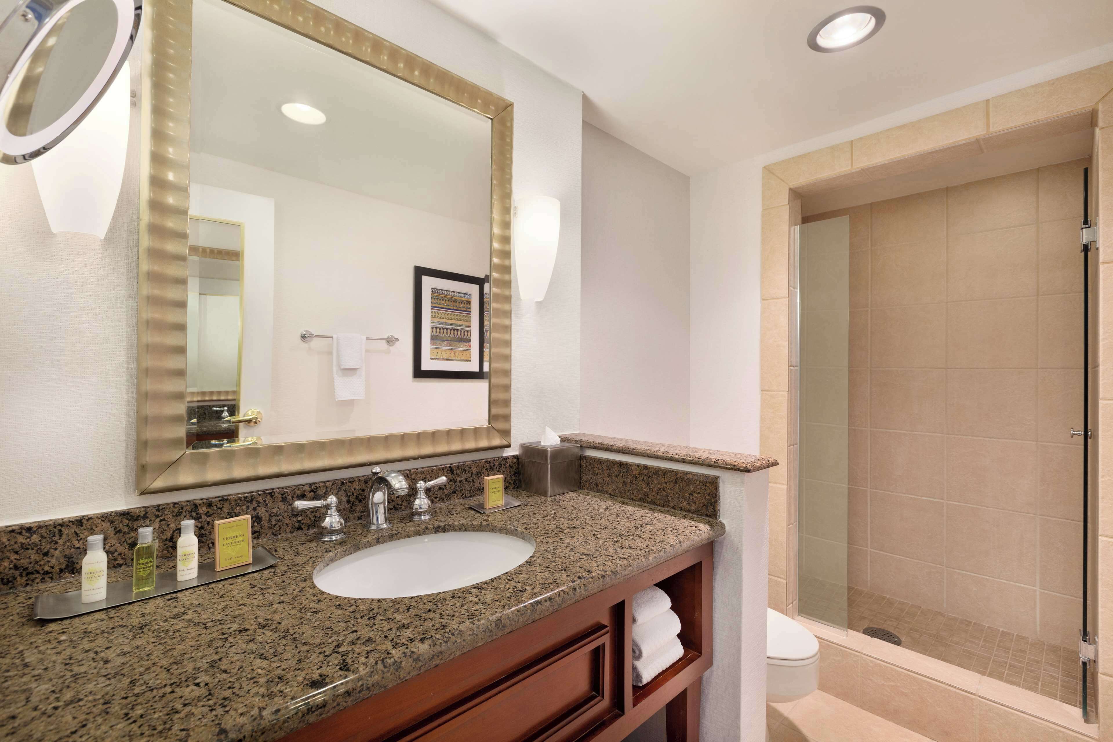 Une salle de bain avec un lavabo doté d’un plan de travail en granit, un grand miroir, un tableau encadré et une douche fermée par des parois de verre.