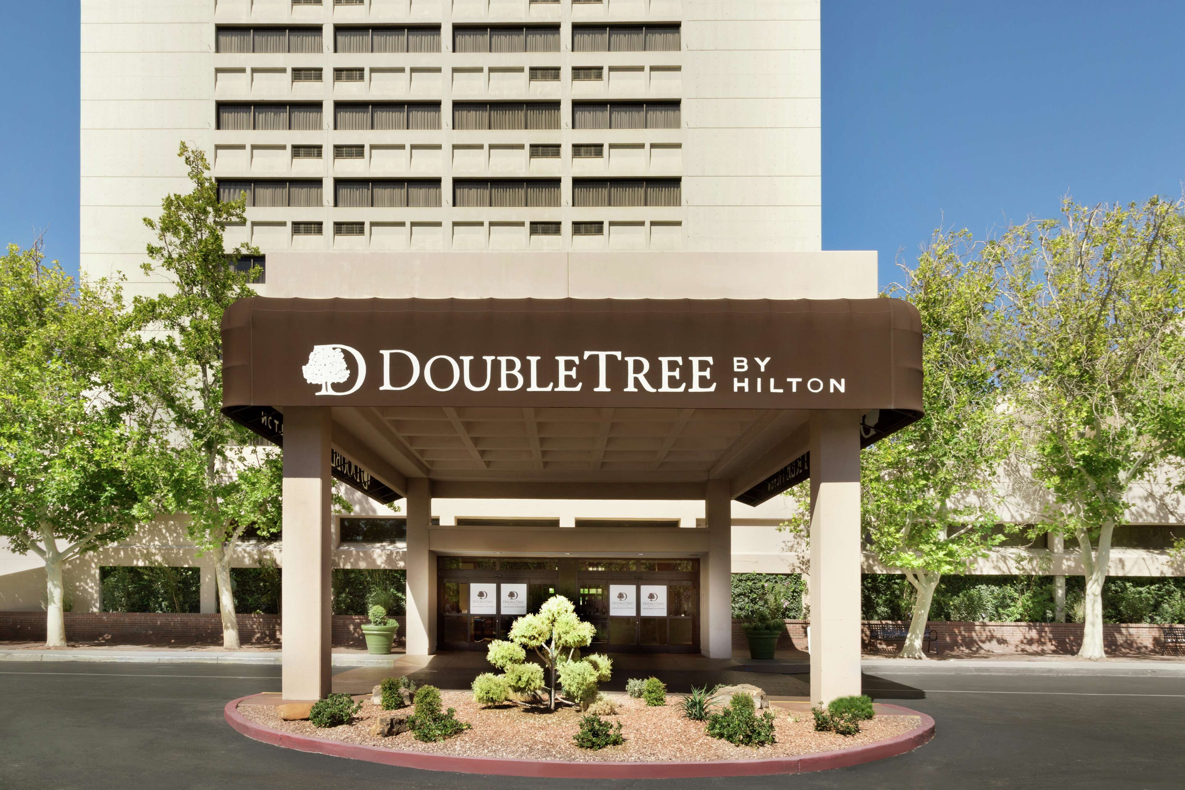 A DoubleTree by Hilton hotel entrance with a covered waiting area, a landscaped garden, and a tall building in the background.
